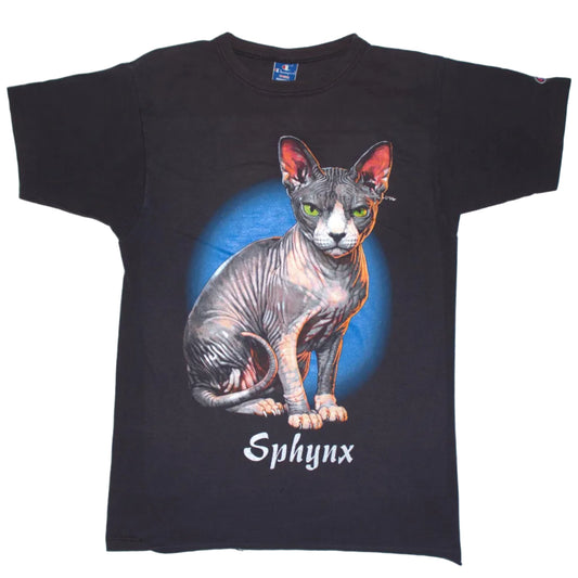A black short-sleeve graphic t-shirt featuring a detailed illustration of a hairless gray and white cat with green eyes sitting in front of a blue circular background, with the word 'Sphynx' printed below in white. The shirt is laid flat against a light gray background.